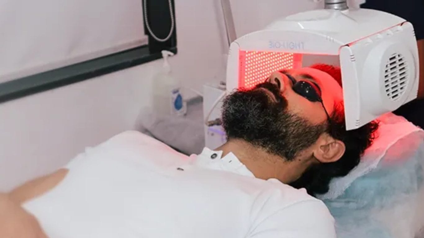 A man lying under a red light therapy panel in a clinic, wearing protective eye shields.